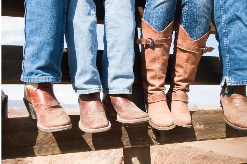 Men wear cowboy boots with jeans and are sitting on the fence at the ranch.