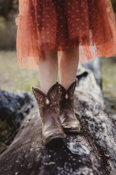 A toddler wears cowboy boots and is walking on the fallen tree