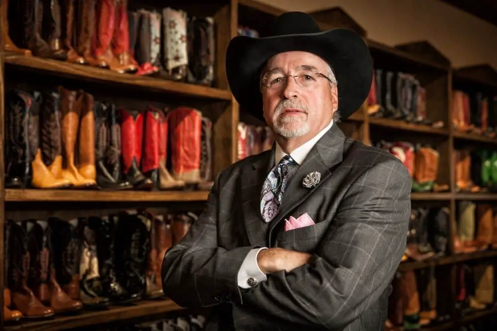 an old business man wearing a cowboy hat and suit standing ahead of a big cowboy boot shelf