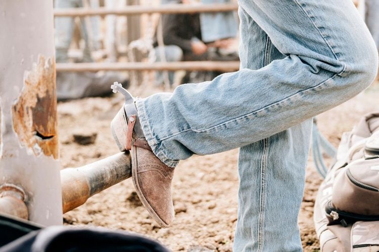 Men wear cowboy boots and jeans standing beside the wooden fence