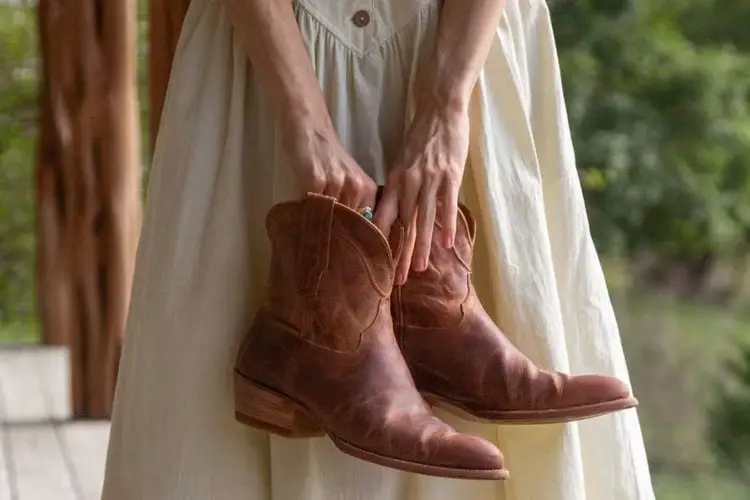 a girl is wearing beige dress and holding a pair of The Penny boots of Tecovas in her hands