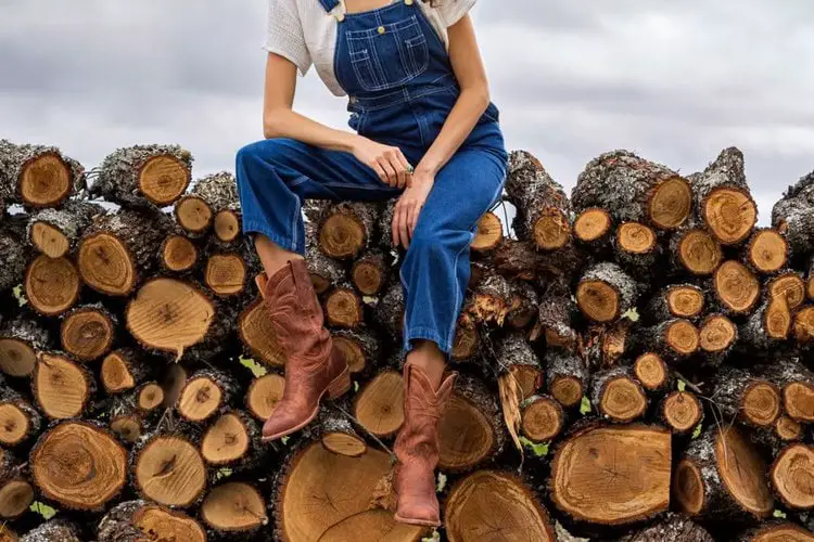 A girl is sitting on stack of woods wearing overalls and The Jamie boots of Tecovas brand