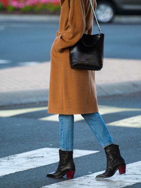 A woman wears black cowboy boots with jeans and jacket and bag and are walking on the street