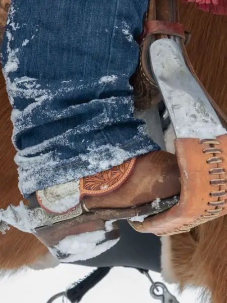 A man wears cowboy boots and are riding a horse under the snow