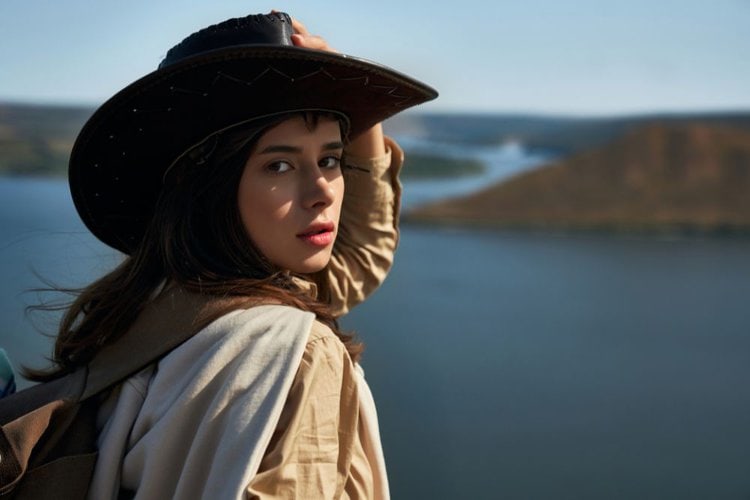 a woman wearing a leather cowboy hat stand near a river