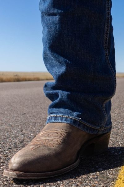 A man on the road under sun wears jeans and cowboy boots