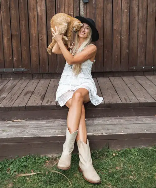 A woman wears white dress with whit cowboy boots and is hugging the dog