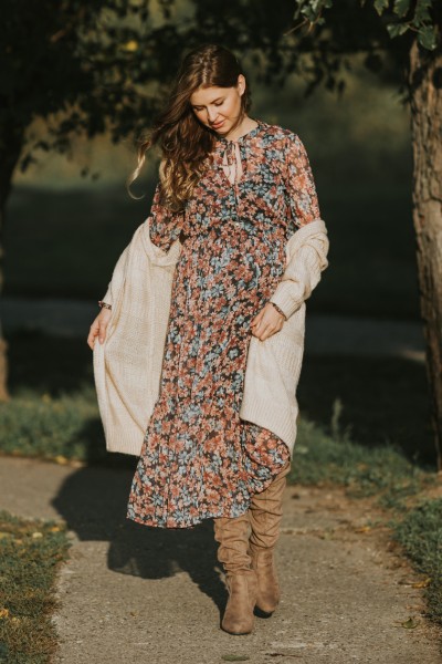 A pregnant woman wears boho dress, cardigan and cowboy boots and is standing on the road