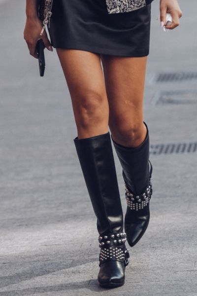 A woman wears black cowboy boots with black mini dress for street style.