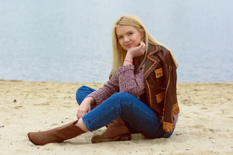 A woman wears jeans with brown cowboy boots, sweater and leather coat