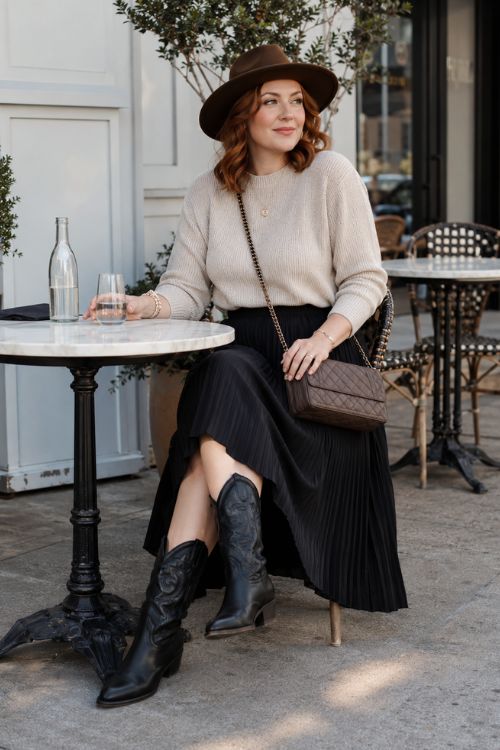 a woman in a black pleated midi skirt, an oatmeal sweater, black cowboy boots, and a felt hat, and a crossbody bag