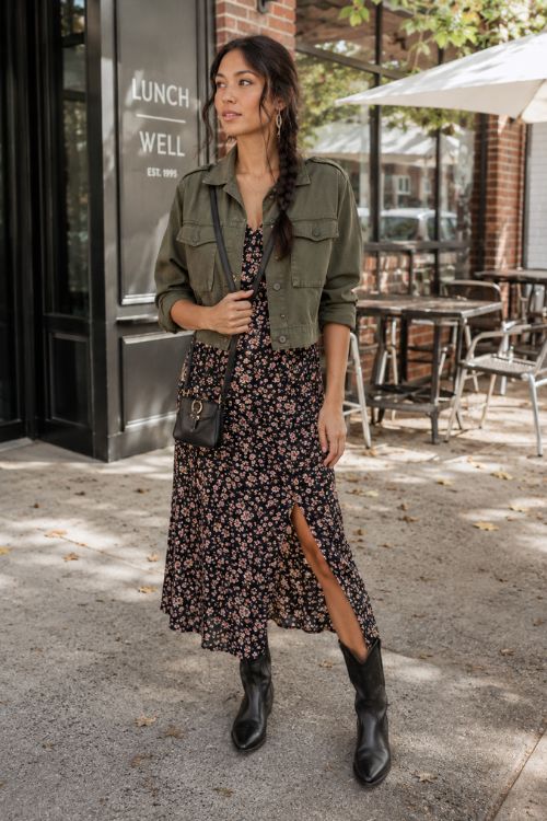 a woman wearing a dark floral midi dress, an olive cropped utility jacket, and black leather cowboy boots
