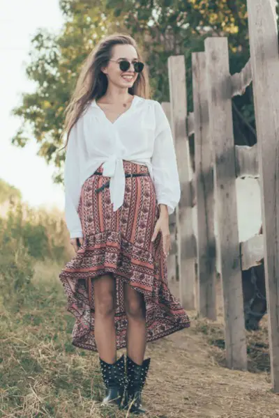 A woman wears a pleated skirt with black cowboy boots and a blouse
