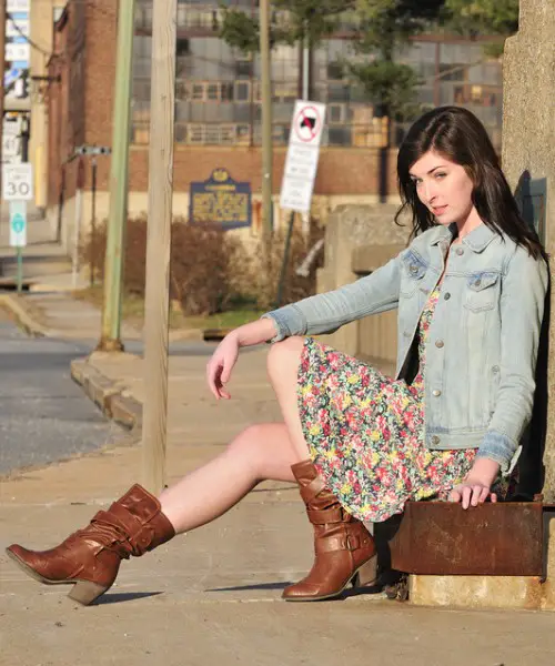 A woman wears cowboy boots, floral dress with denim jacket