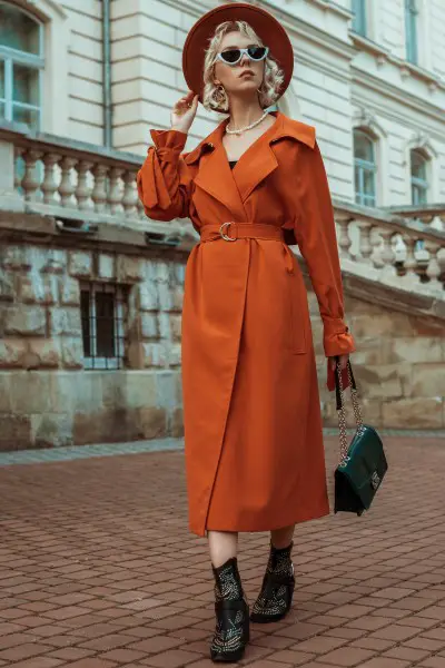 A woman wears orange trench coat with cowboy boots and wide brim hat