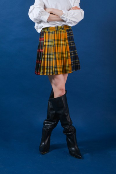 A woman wears plaid pleated skirt with a blouse and black cowboy boots on a blue background