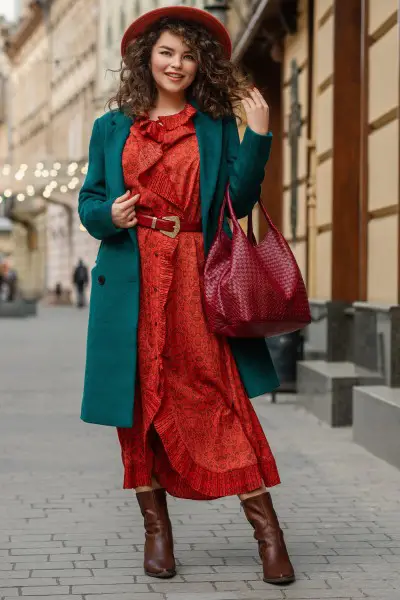 A woman wears red dresses with brown cowboy boots, green blazer and a tote bag.