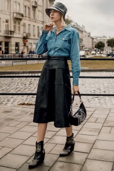 A woman wears black ankle cowboy boots with black leather midi skirt and denim shirt