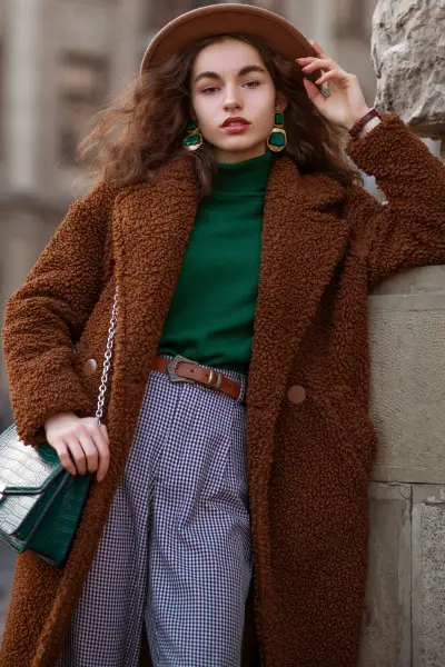 A woman wears brown cowbo boots, fur coat with purpose pants and green top