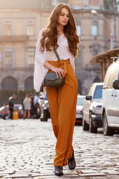 A woman wears yellow high-waisted wide-leg pants, white blouse, ankle cowboy boots and holding green leather bag.
