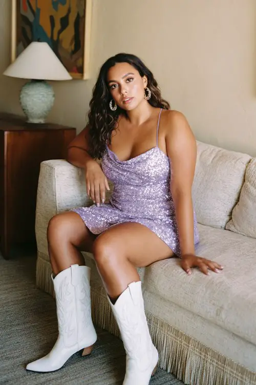 A curvy woman wears a glittery lavender sequin dress with white cowboy boots and hoop earrings at a girls-only party