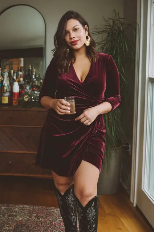 A curvy woman wears a velvet burgundy wrap dress, black cowboy boots with embroidery, and gold hoop earrings