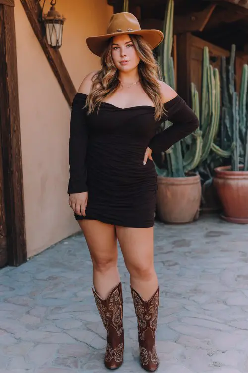 A plus-size woman wears a black off-shoulder mini dress, chocolate brown cowboy boots with embroidered details, and a wide-brim hat