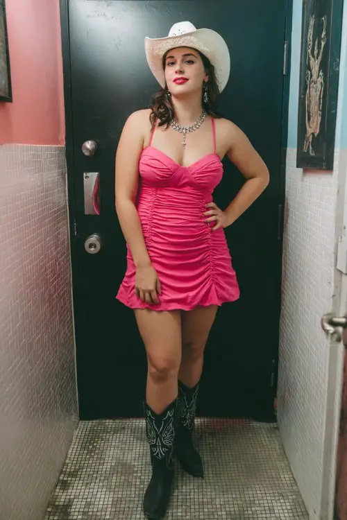 A plus-size woman wears a ruched hot pink dress with rhinestone-embellished black cowboy boots and a white cowgirl hat