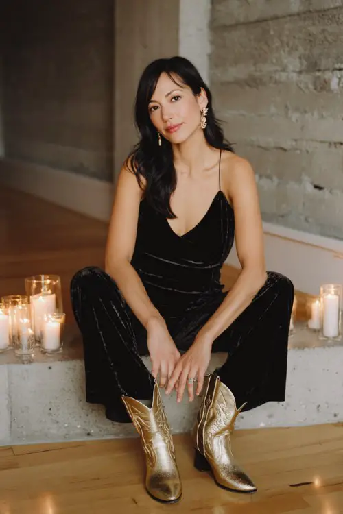 A woman over 30 wears a black velvet jumpsuit with a plunging neckline and gold cowboy boots