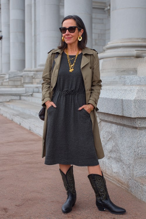 A woman over 40 wears black cowboy boots with a charcoal midi knit dress, gold hoop earrings, and a wool trench
