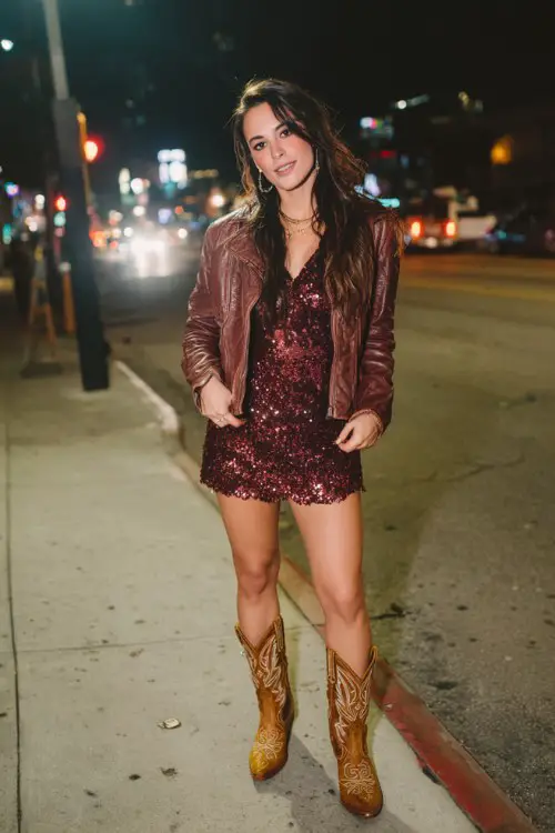 A woman styles brown cowboy boots with a sequined burgundy mini dress and a cropped leather jacket