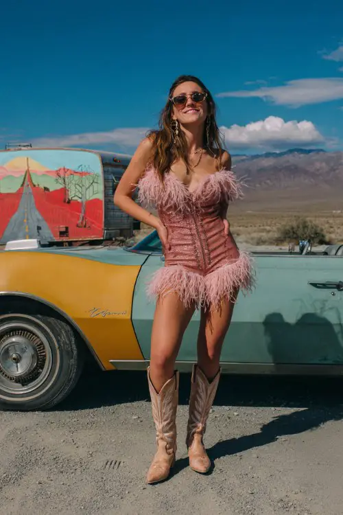 A woman wears a feather-trimmed mini romper in rose gold with matching cowboy boots