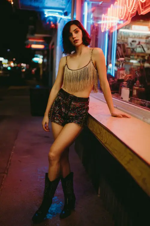 A woman wears a fringe crop top with sequin shorts and black cowboy boots, standing by a neon-lit bar