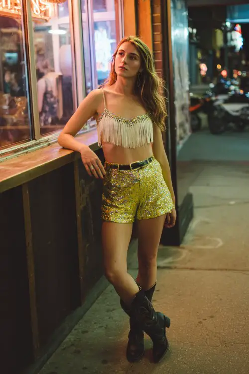 A woman wears a fringe crop top with sequin shorts and black cowboy boots, standing by a neon-lit bar