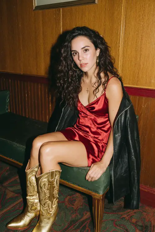 A woman wears a red satin slip dress with a black leather jacket and gold cowboy boots