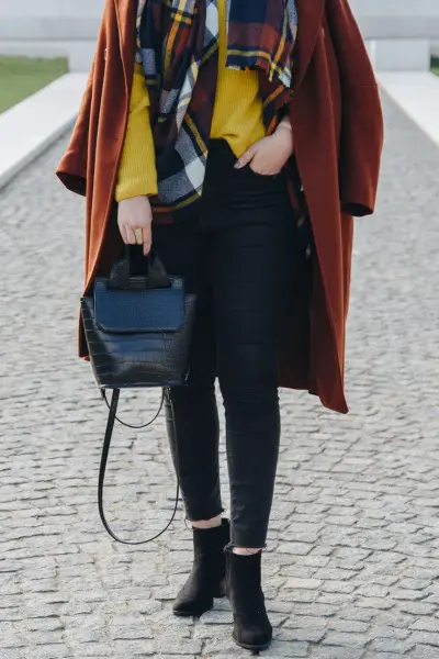A woman wears black ankle boots, black jeans, yellow sweater, dark red trenchcoat, and black handbag