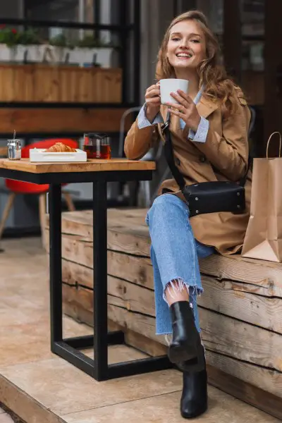 A woman wears black ankle boots with jeans and trench coat at the coffee shop