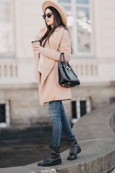 A woman wears long coat with jeans, black boots and wide brim hat