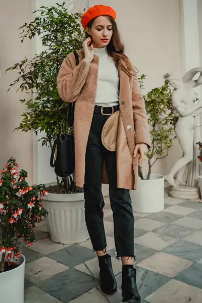 A woman wears short cowboy boots, black jeans, trench coat, white turtleneck and beret.