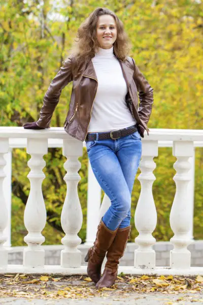 A woman wears skinny jeans, cowboy boots, white turtleneck and leather coat