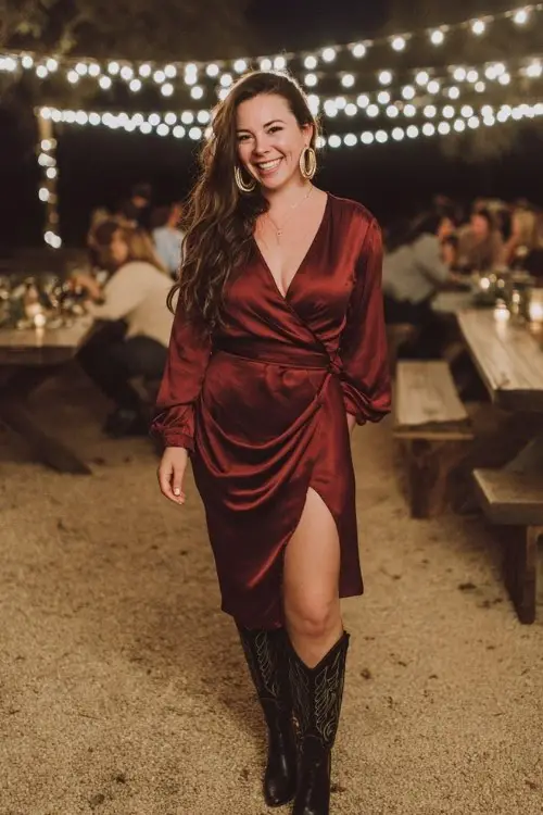 A curvy woman wears a burgundy satin wrap dress with a slit, styled with tall black cowboy boots and gold hoop earrings