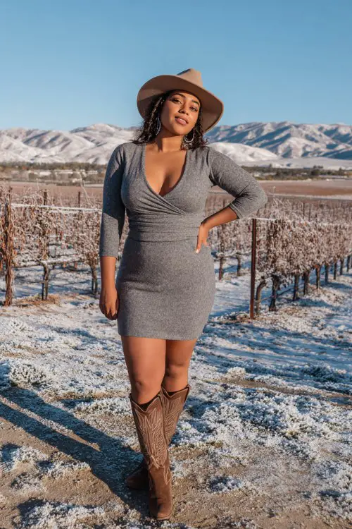 A curvy woman wears a gray wool mini dress with a fitted waist, styled with tall brown cowboy boots and a matching felt hat