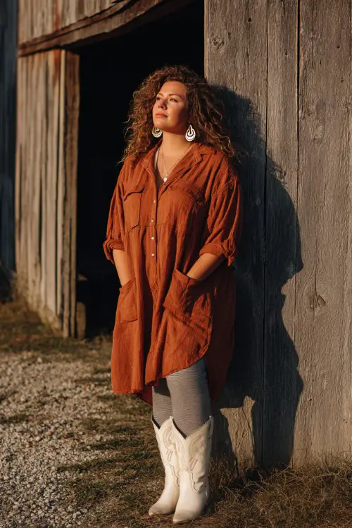 A curvy woman wears a loose tunic dress over gray leggings with white cowboy boots, styled with statement earrings