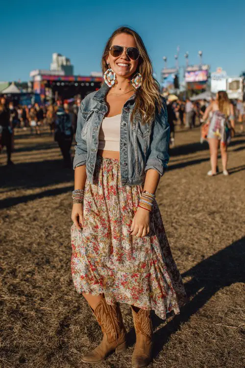 A curvy woman wears brown cowgirl boots with a flowy floral midi skirt and cropped denim jacket, accessorized with hoop earrings and stacked bracelets