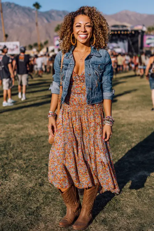 A curvy woman wears brown cowgirl boots with a flowy floral midi skirt and cropped denim jacket, accessorized with hoop earrings and stacked bracelets