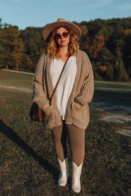 A curvy woman wears ribbed leggings with cream short cowboy boots, a chunky oversized cardigan layered over a simple tee, felt hat and crossbody bag finishing the outfit