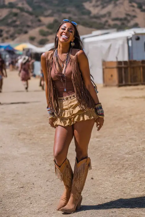 A curvy woman wears tan cowgirl boots with a ruffled boho mini skirt and fitted knit top, layered with a fringe vest and stacked bracelets