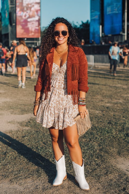 A curvy woman wears white ankle cowboy boots with a ruffled boho mini dress layered under a cropped suede jacket, accessorized with stacked bracelets and a fringe clutch