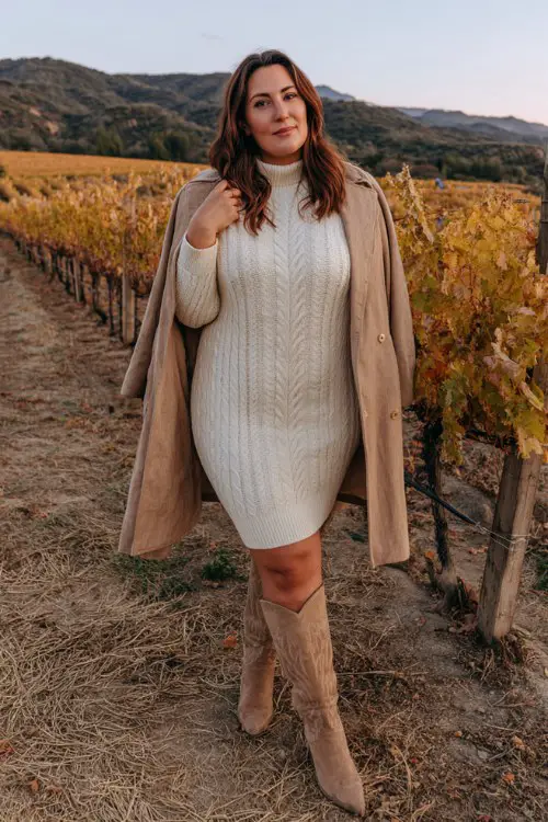 A plus-size woman wears a cream knit dress with a cable texture and mock neck, styled with tall suede cowboy boots and a wool coat
