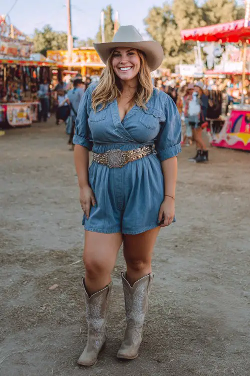 A plus-size woman wears a denim romper cinched with a concho belt, styled with tall embroidered cowboy boots and a felt hat